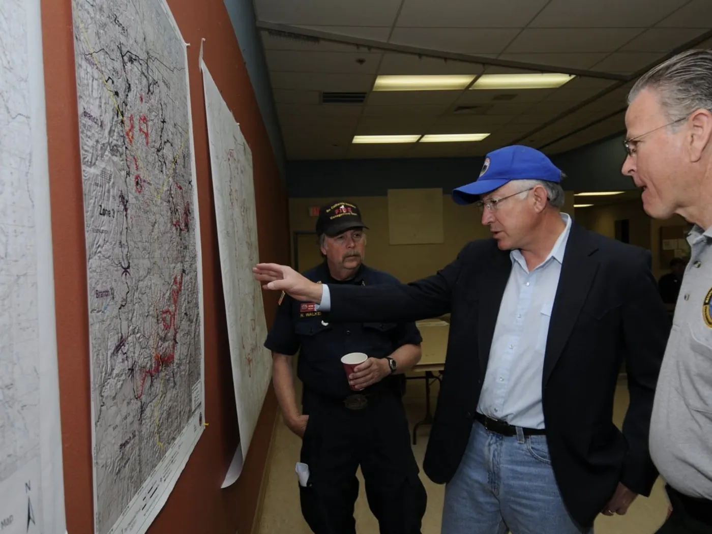 Ken Salazar pointing to a wildfire map on the wall while firefighters look on