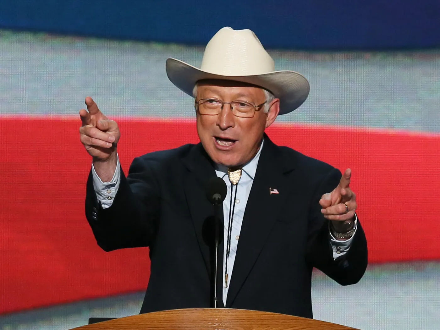 Ken Salazar in his cowboy hat at a podium, pointing out to the crowd with both hands, a flag backdrop behind him