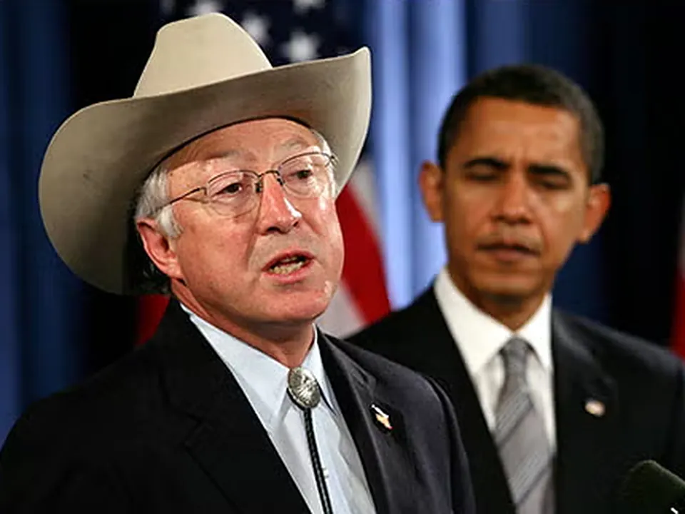 Ken Salazar in his hat, speaking at a bank of microphones with Barack Obama standing behind him