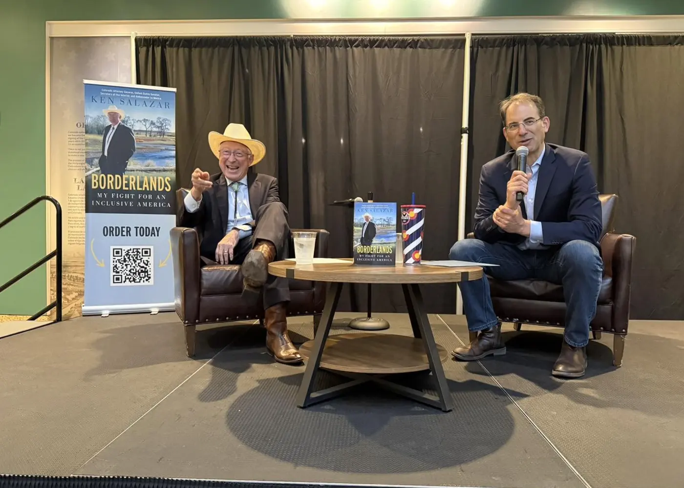 Ken Salazar laughing and pointing to the audience during an on-stage conversation, a Borderlands banner beside him